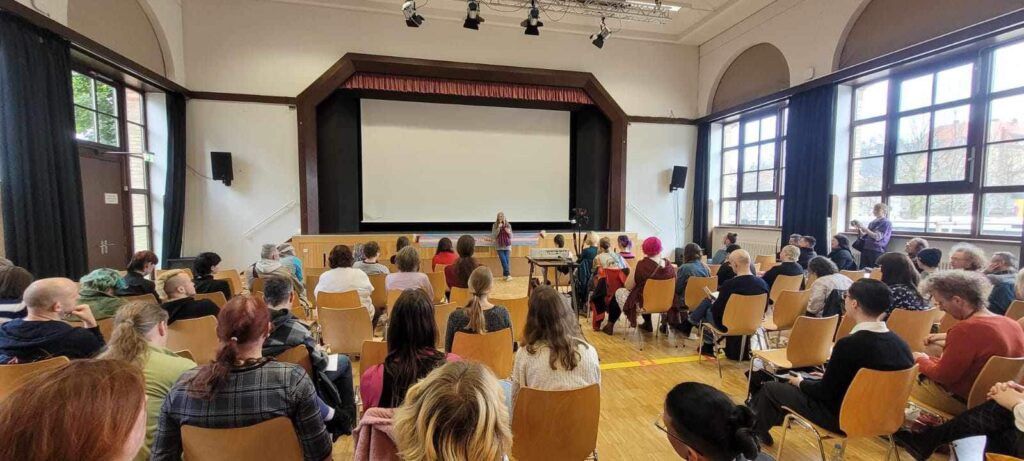 Foto vom Seminarraum im Alten Feuerwehrhaus Stuttgart. Auf vielen Stühlen sitzen Menschen im Publikum.